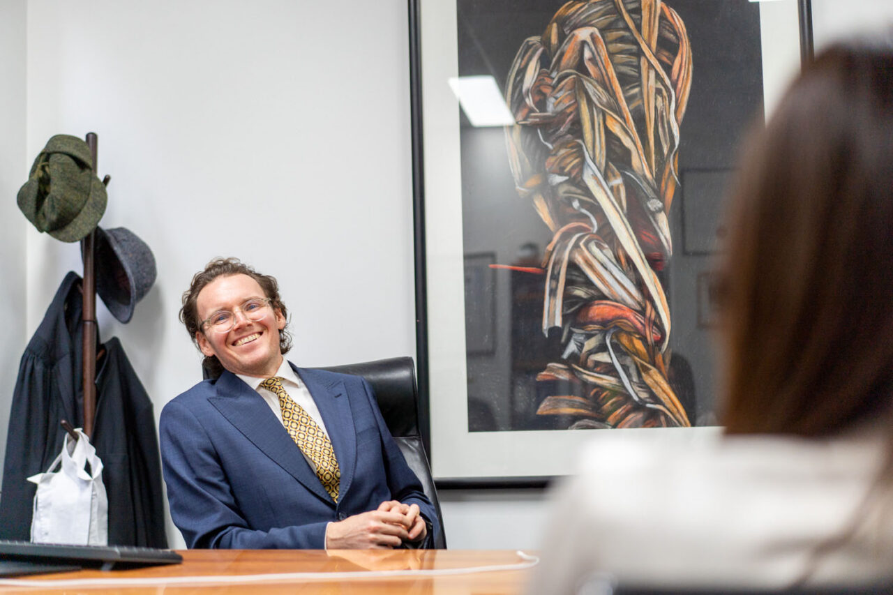 Male barrister sitting at a timber desk smiling during a documentary brand photography session in a Melbourne legal office, large artwork on the wall behind him