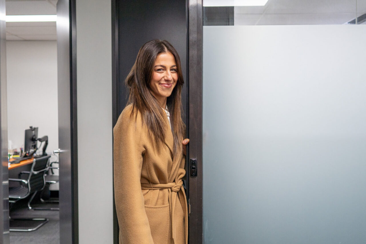 Female lawyer standing in a doorway of a Melbourne legal office smiling over her shoulder, wearing a camel coat with frosted glass partitions and office in the background