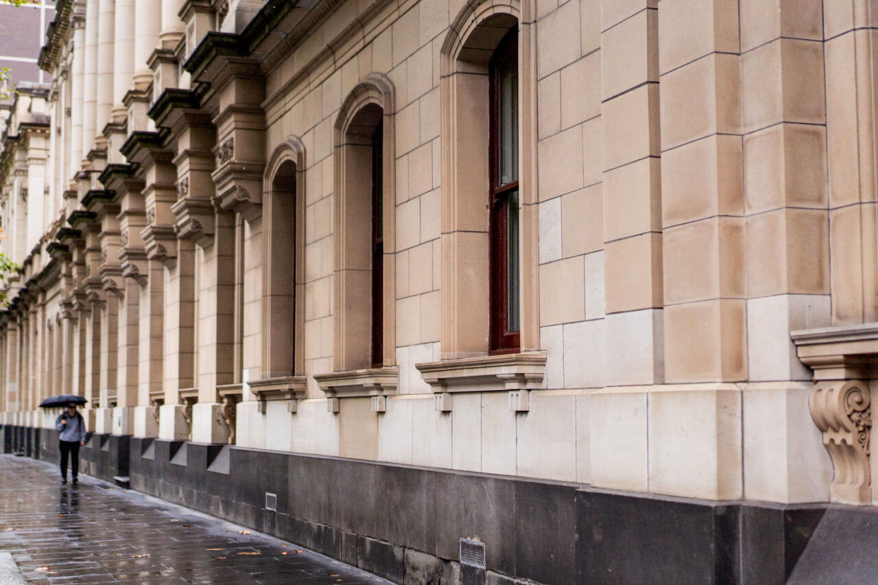 Heritage sandstone facade of the Melbourne Law Courts building on a wet day with a lone figure walking past under an umbrella