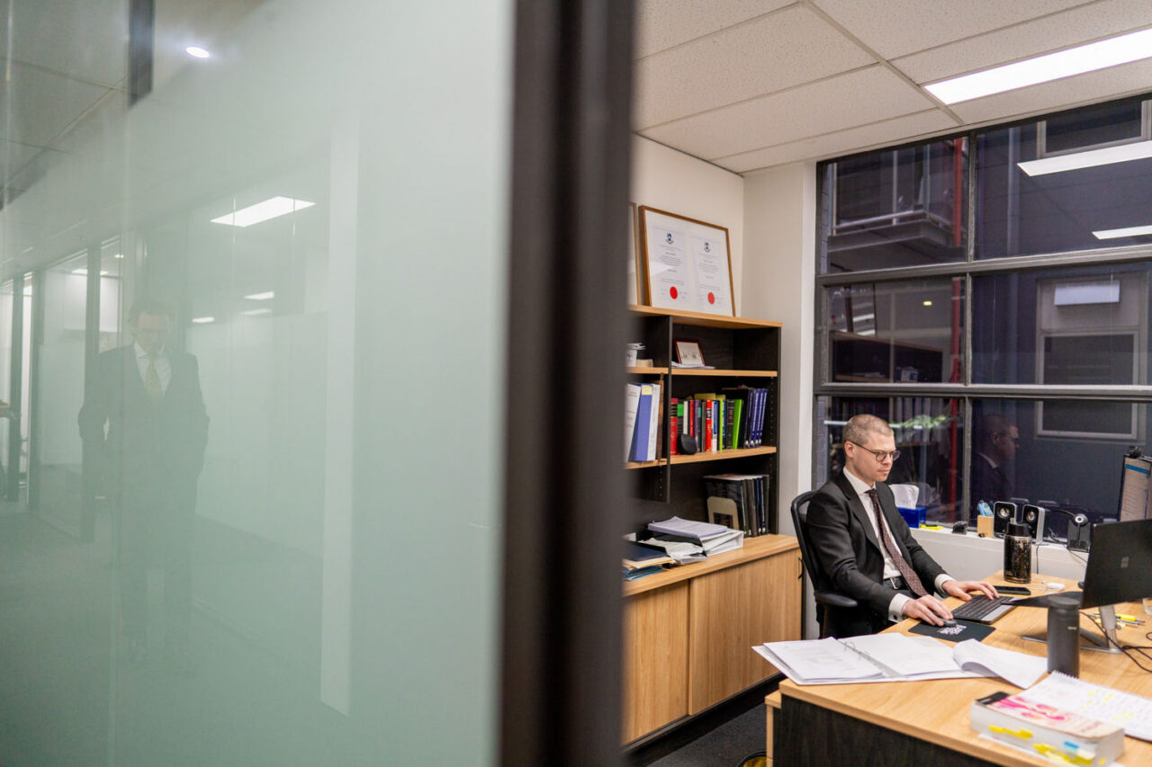 Male lawyer working at his desk in a glass-walled Melbourne office, framed certificates on the bookshelf behind him, reflected in the frosted glass partition