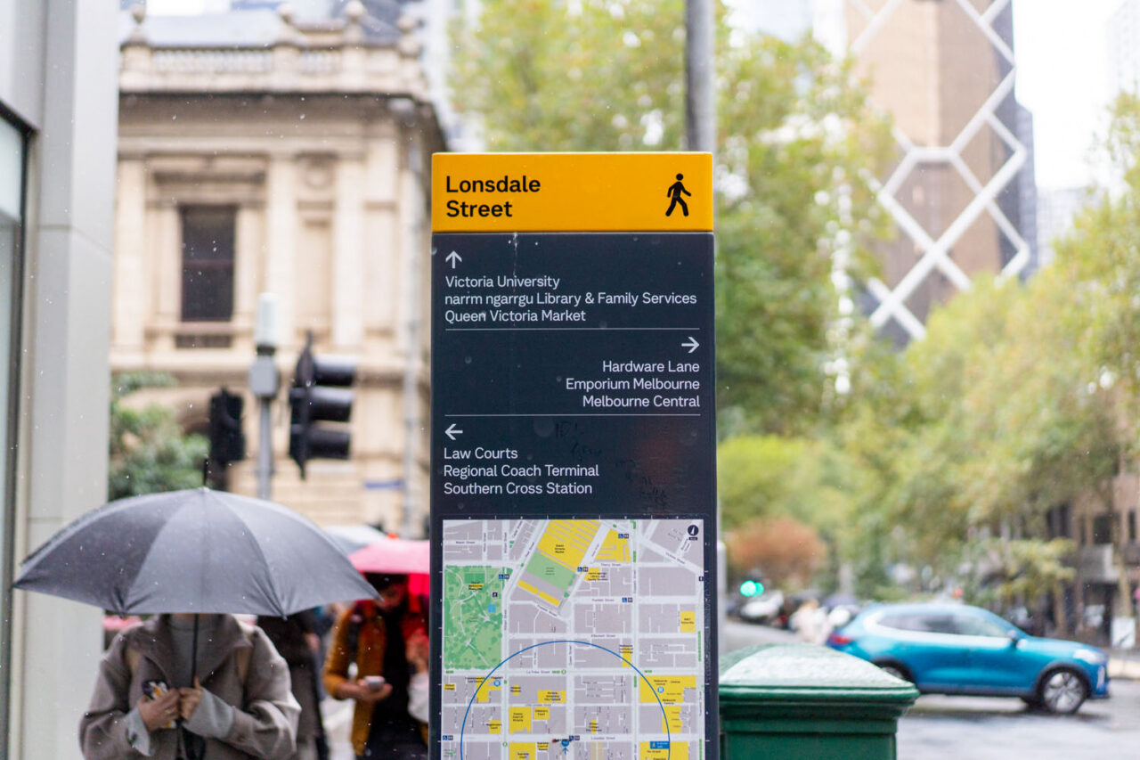 Lonsdale Street pedestrian wayfinding sign in Melbourne CBD on a rainy day with a person holding an umbrella and heritage buildings in the background