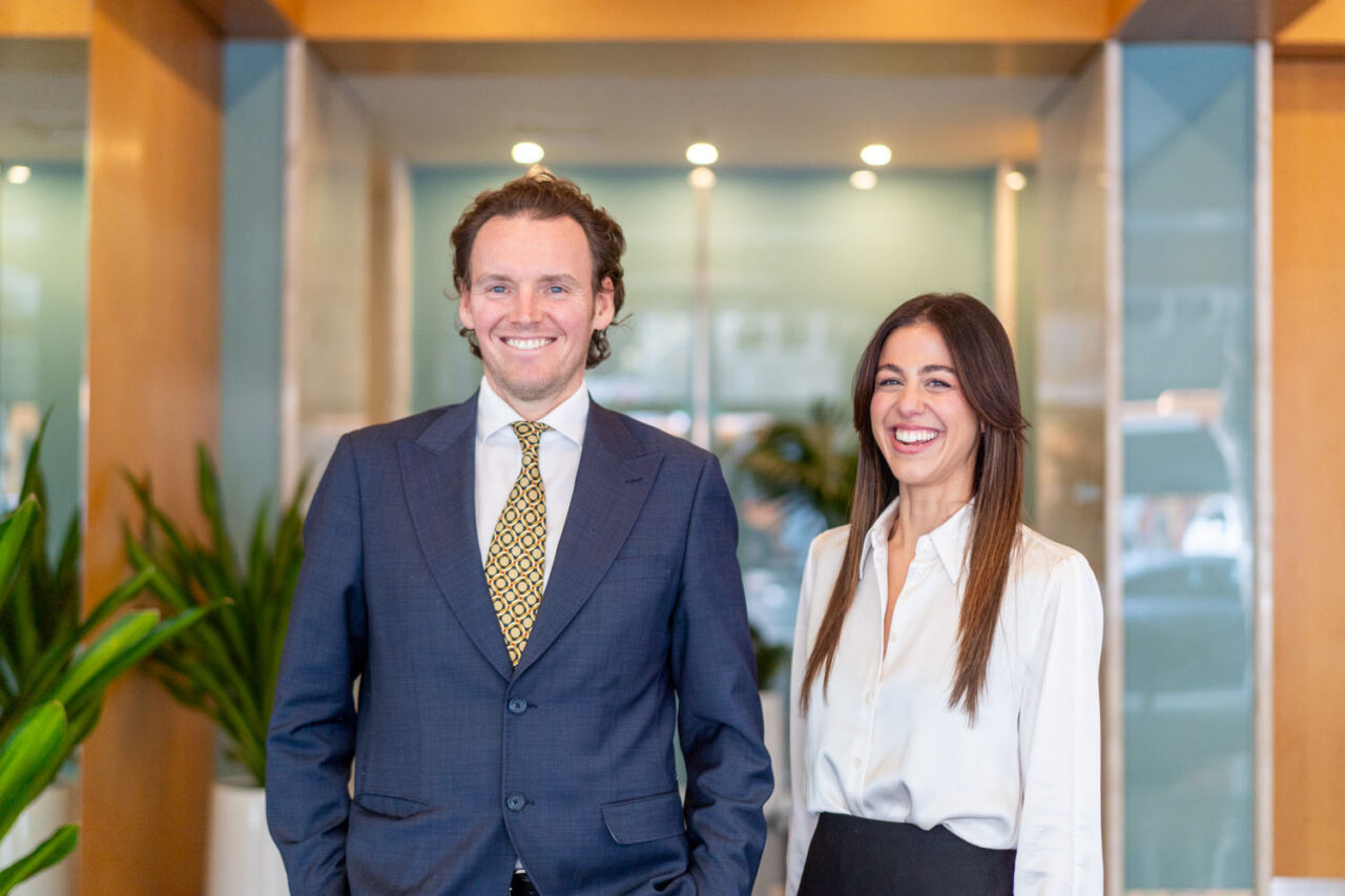 Male and female lawyer standing together smiling in the foyer of a Melbourne legal office, warm timber and glass walls in the background