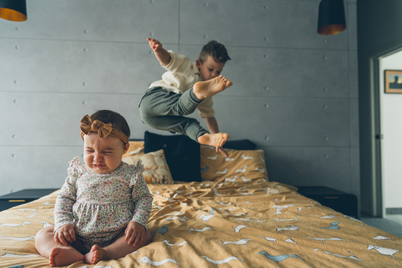 A baby sitting on a mustard yellow bed looking unimpressed while her older sibling leaps through the air behind her during an in-home documentary family session