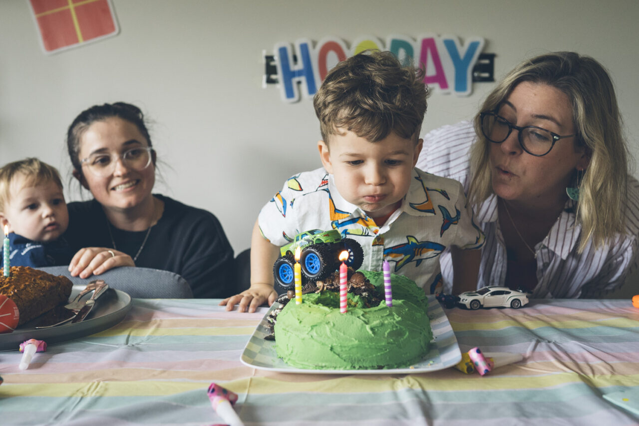 Toddler blowing out birthday candles on a green monster truck cake, documentary family home photography Melbourne