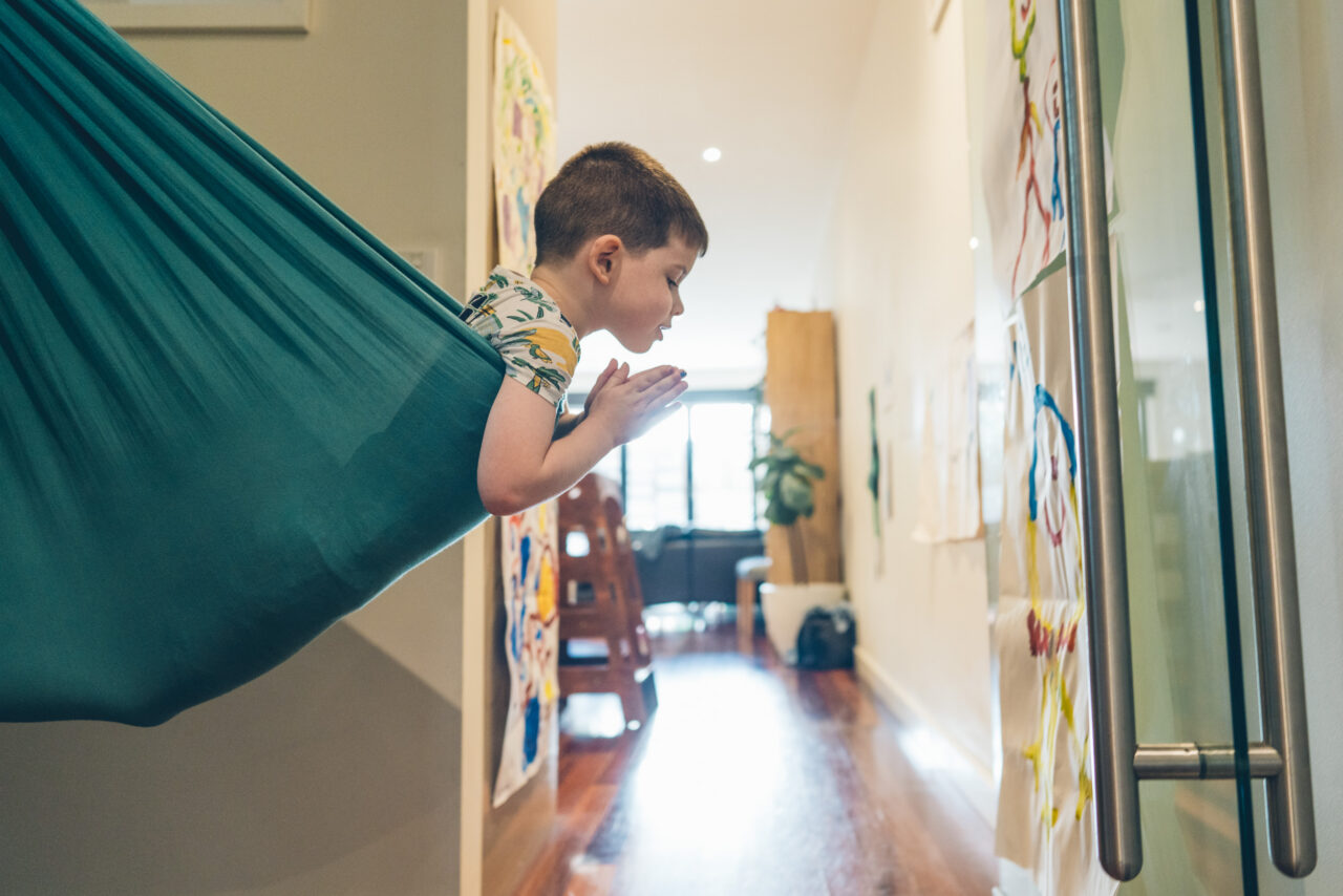 A young boy leans out of a teal indoor hammock hung in a hallway lined with children's artwork, absorbed in quiet thought during an in-home documentary family photography session