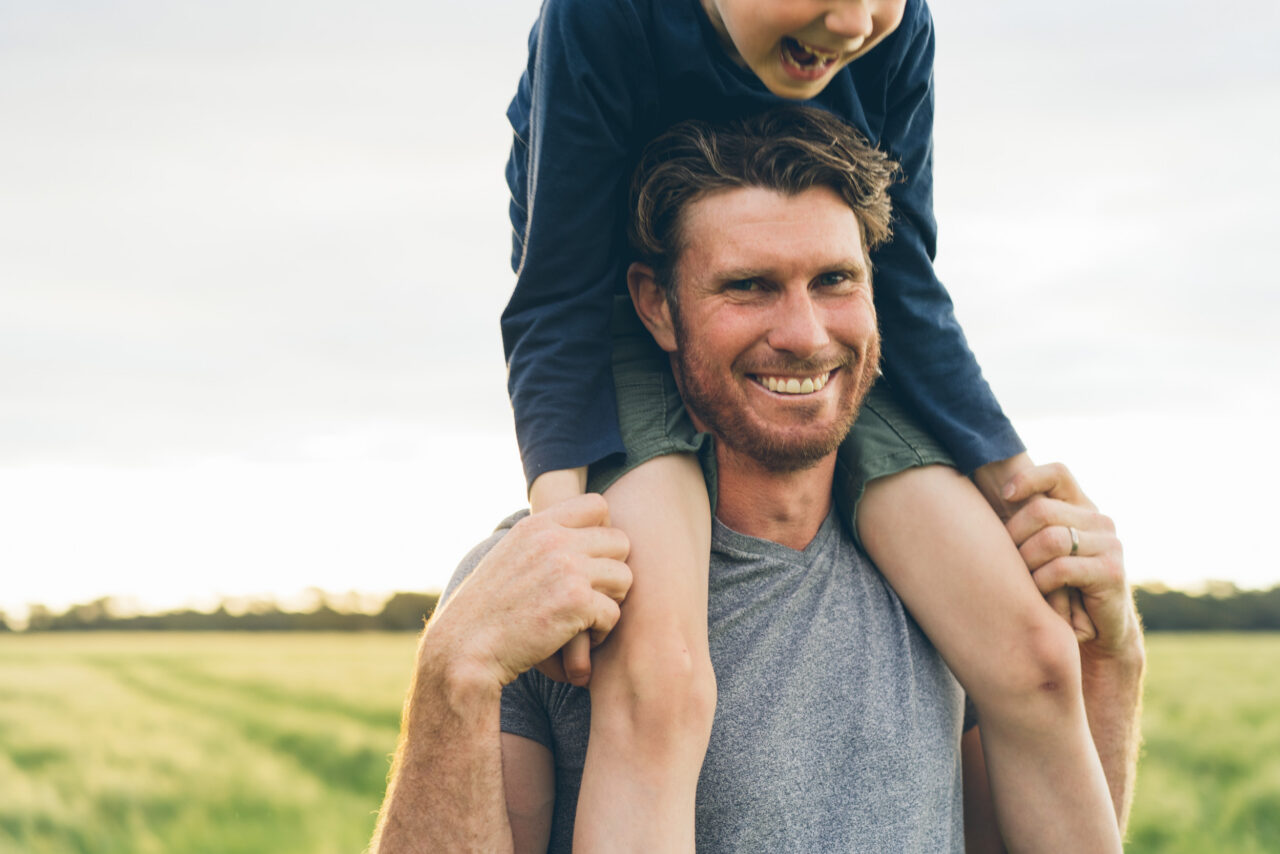 father carries his young son on his shoulders in a green paddock at golden hour, both smiling, captured during a documentary family photography session on the Surf Coast