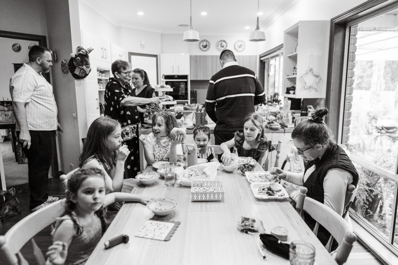 Black and white photograph of a large extended family gathering around a long dining table, multiple children eating lunch while adults move around the busy kitchen behind them during a documentary family photography session