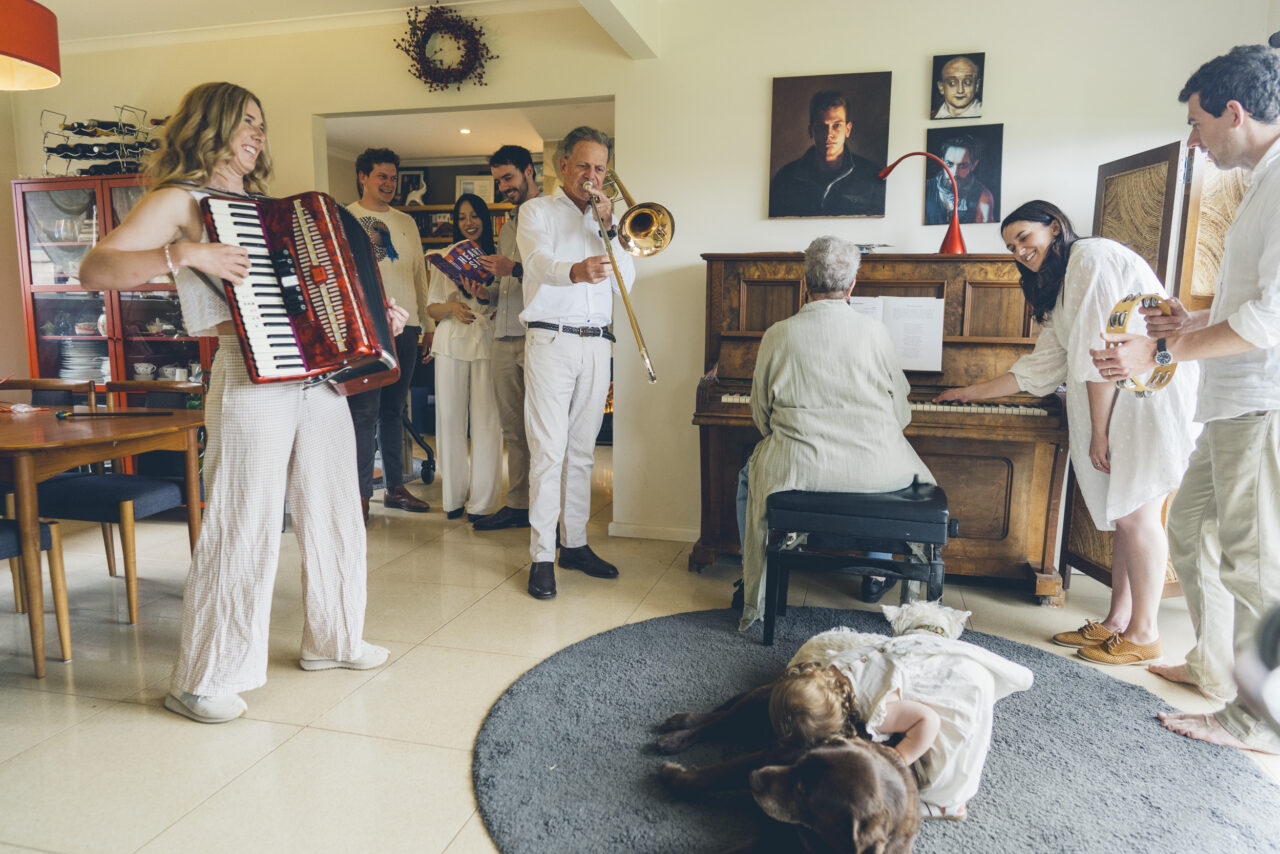 An extended family gathered in a living room playing instruments together including accordion, trombone and piano, with a toddler on the floor hugging the dog, during an in-home documentary Your Story photography session
