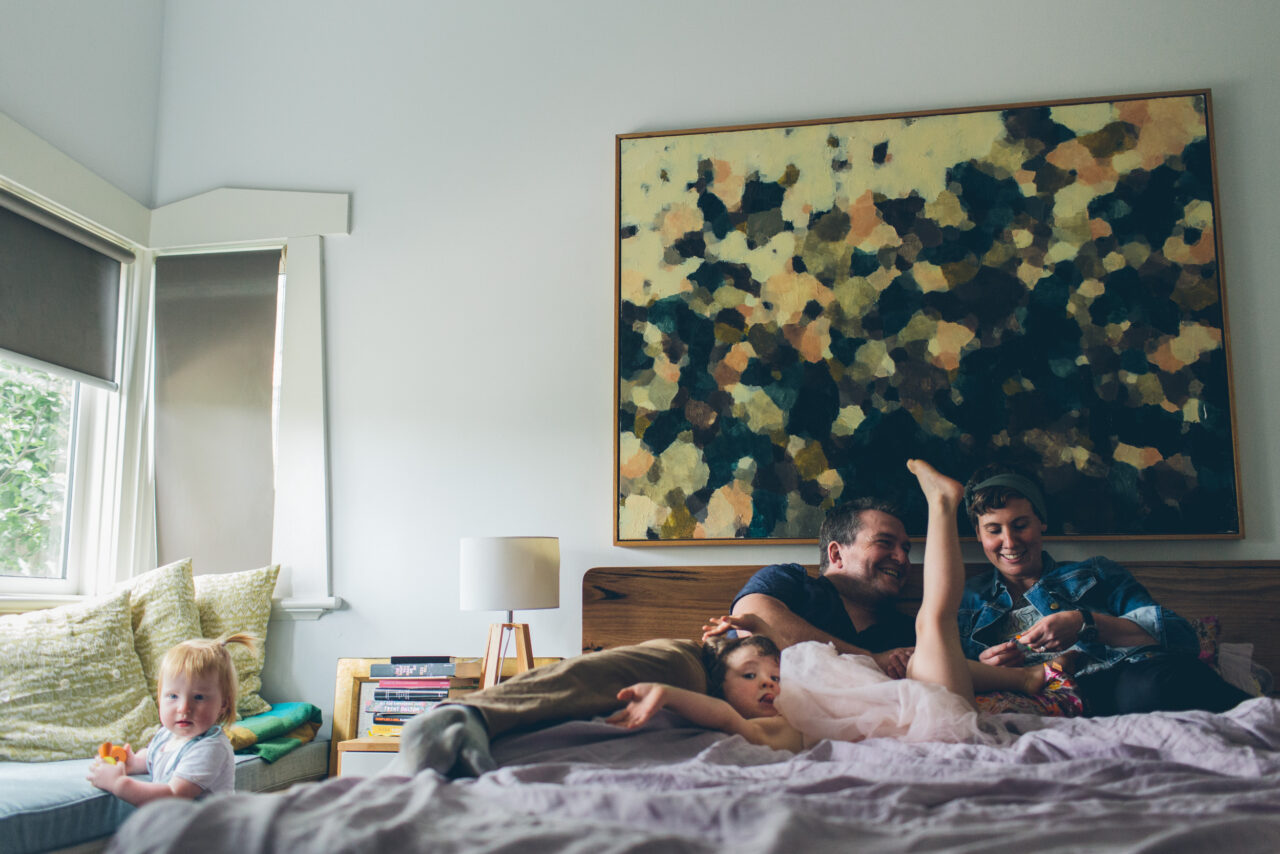 Family of four relaxing on a bed together beneath a large abstract painting, documentary family home photography Melbourne