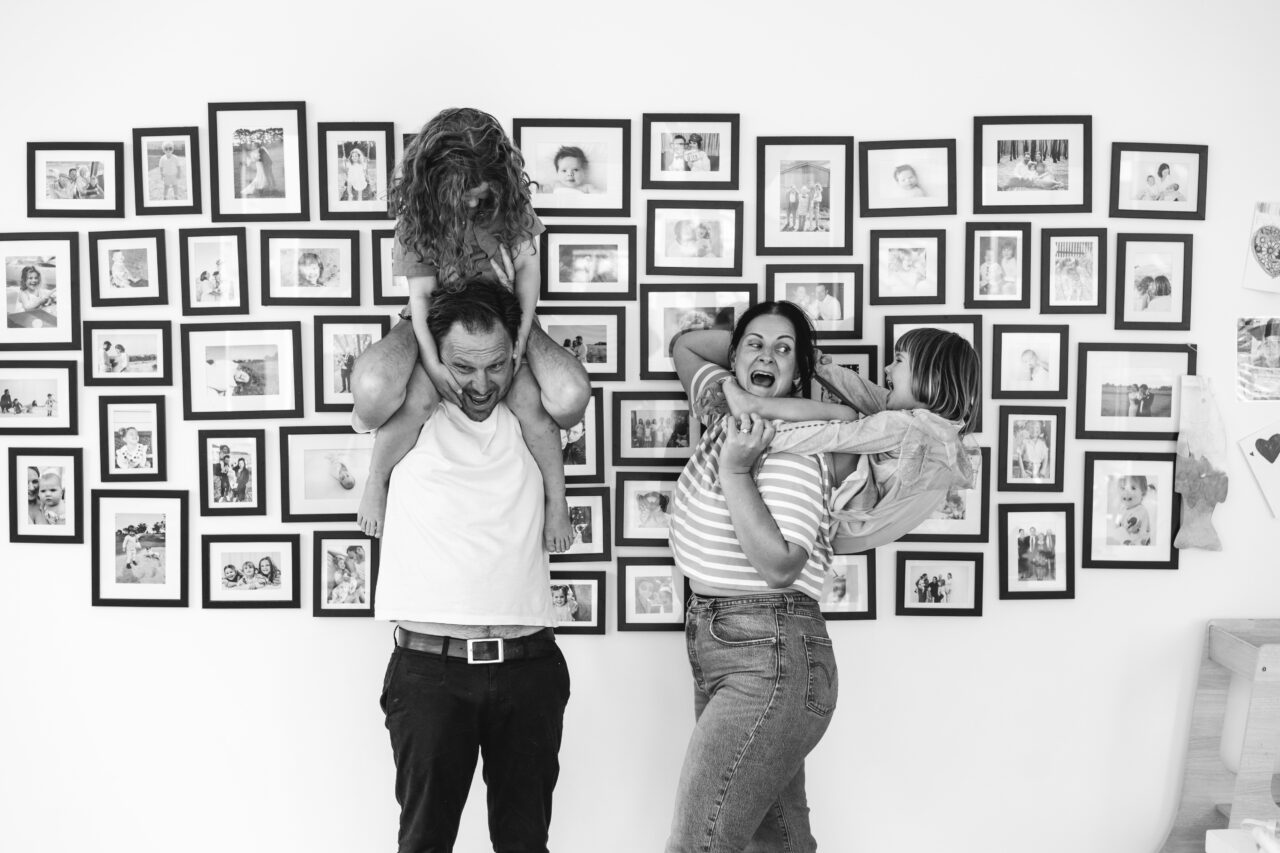 Family of four in front of a large gallery wall of photographs, children climbing on parents, black and white documentary family home photography