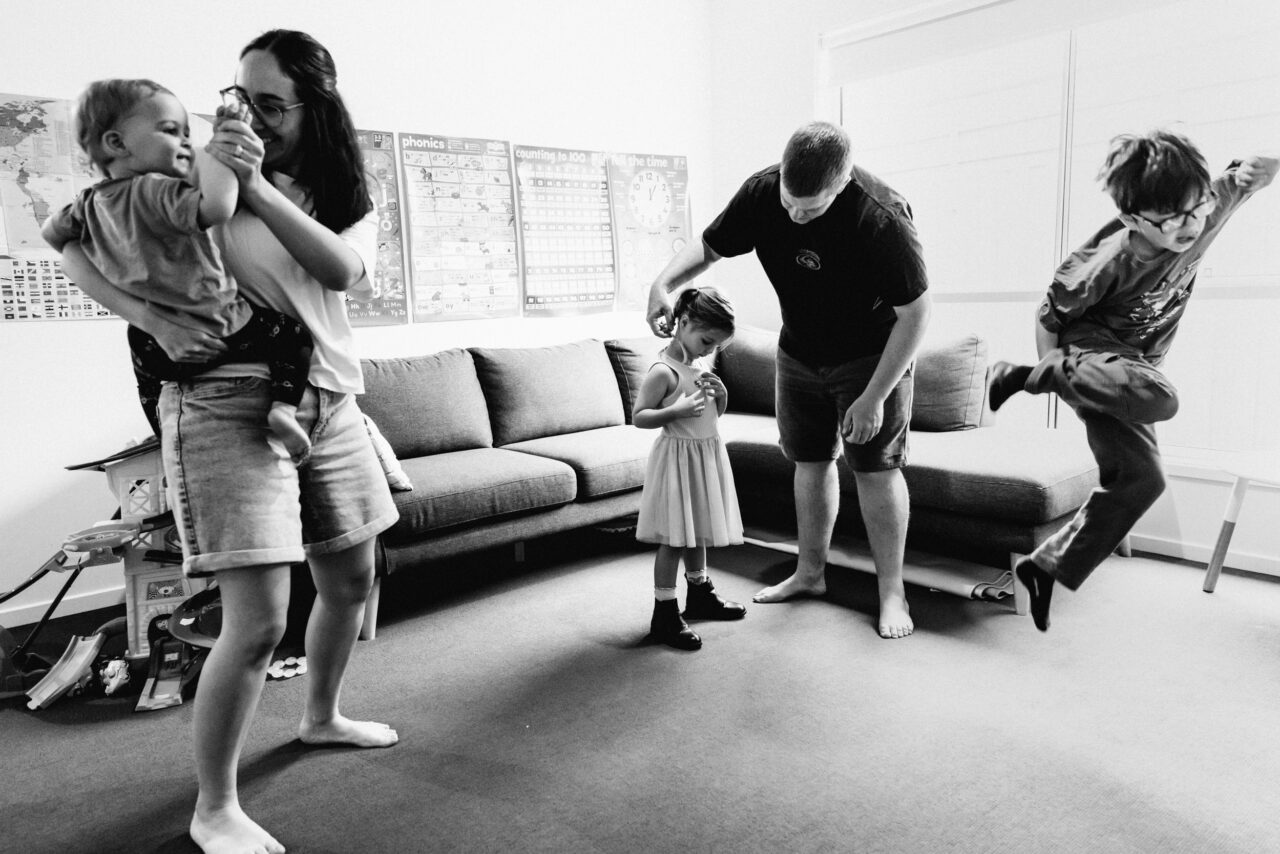 Black and white documentary photograph of a busy family living room, mum holding a baby, dad doing a daughter's hair, and an older child leaping through the air, phonics charts on the wall behind them