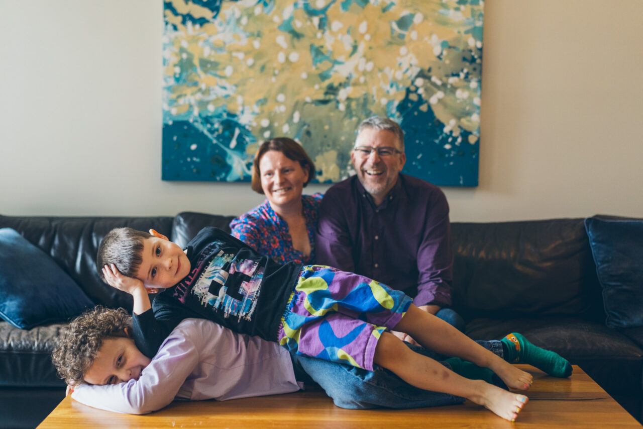 A family of four on a leather couch, two parents laughing behind their two boys who are sprawled across each other and the coffee table, large abstract artwork on the wall behind them