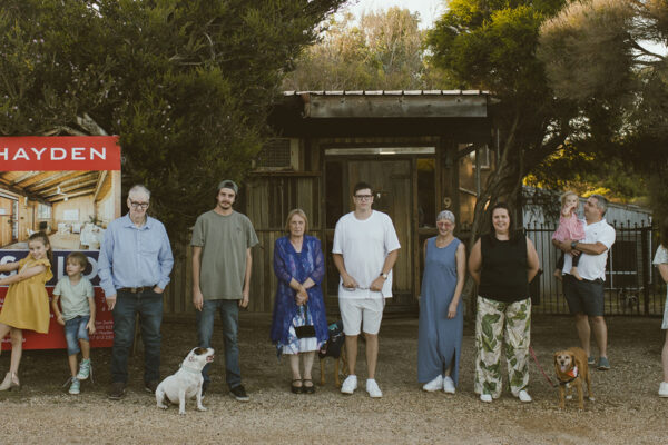 An extended family of three generations lined up outside their sold Jan Juc home on the Surf Coast, two dogs included, in a wide panoramic documentary photograph