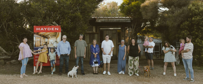 An extended family of three generations lined up outside their sold Jan Juc home on the Surf Coast, two dogs included, in a wide panoramic documentary photograph