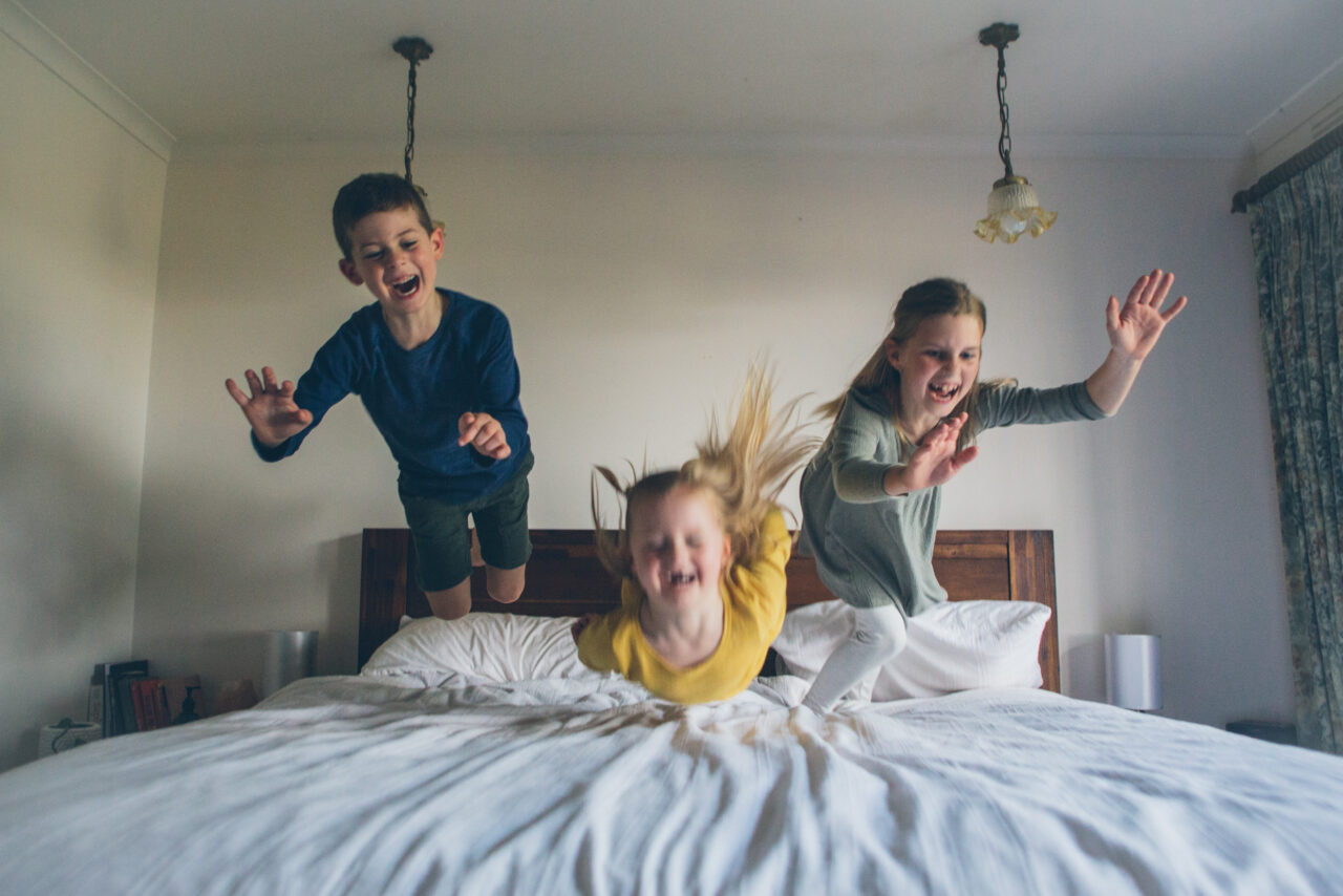 Three children mid-air jumping on a bed, laughing and screaming, captured during an in-home documentary family photography session on the Surf Coast