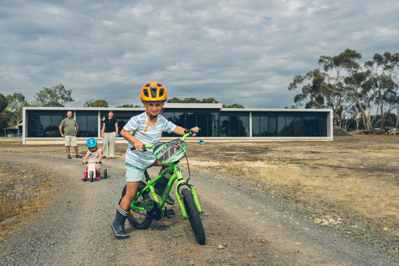 A young boy on a green bike with an orange helmet in the foreground, a toddler on a pink bike behind him, parents watching from in front of a long modern home under a dramatic cloudy sky in regional Victoria