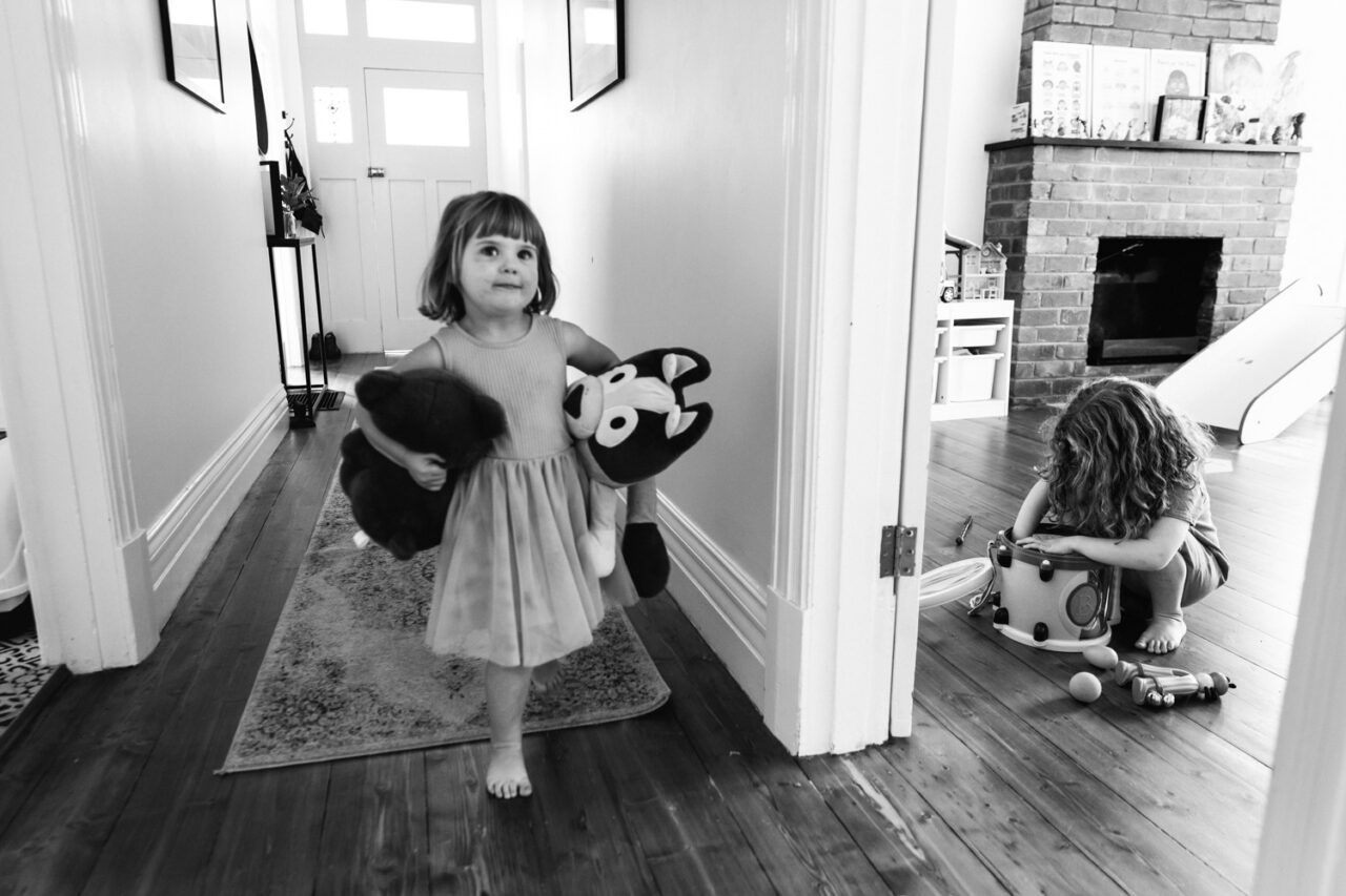 A young girl walks through a hallway carrying two large stuffed animals with purpose, while her sibling plays with toys by the fireplace behind her, captured in black and white during a documentary in-home family session