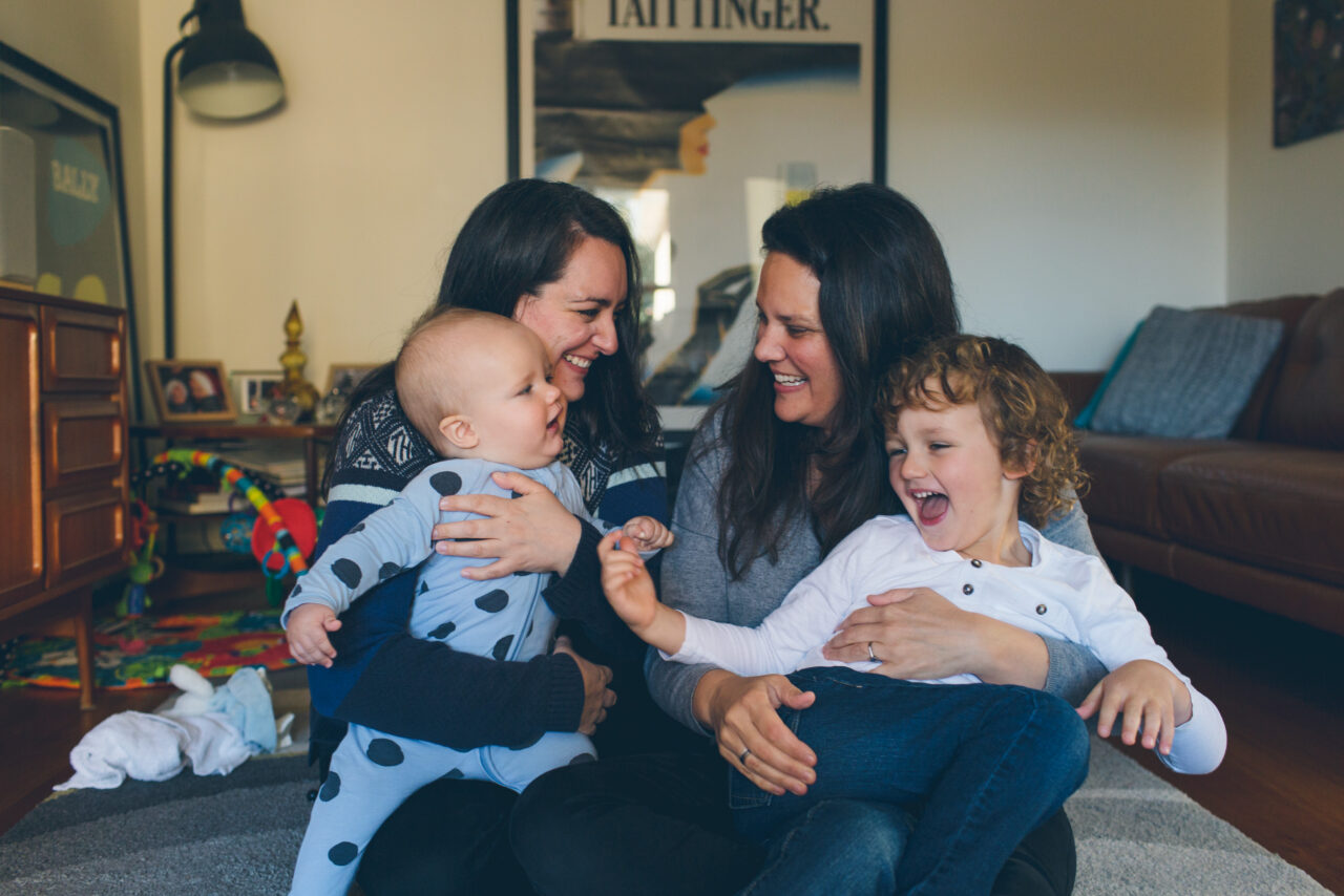 Two mothers sitting on the floor laughing with their babies in a family home lounge room, documentary family home photography Melbourne