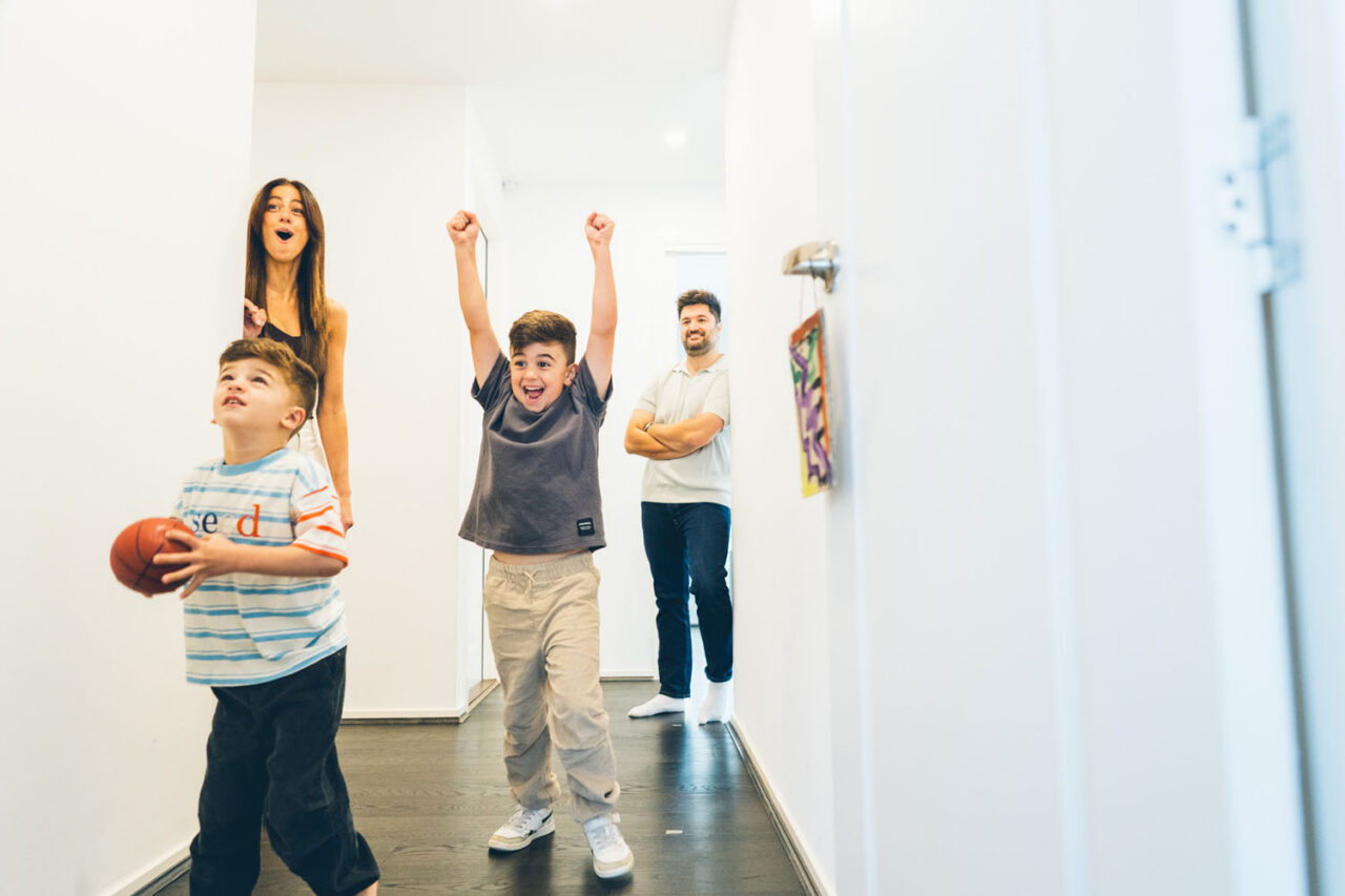 Two boys playing basketball in the hallway during an in-home documentary family session in Melbourne, older boy celebrating with arms raised while younger boy holds the ball and parents watch laughing in the background