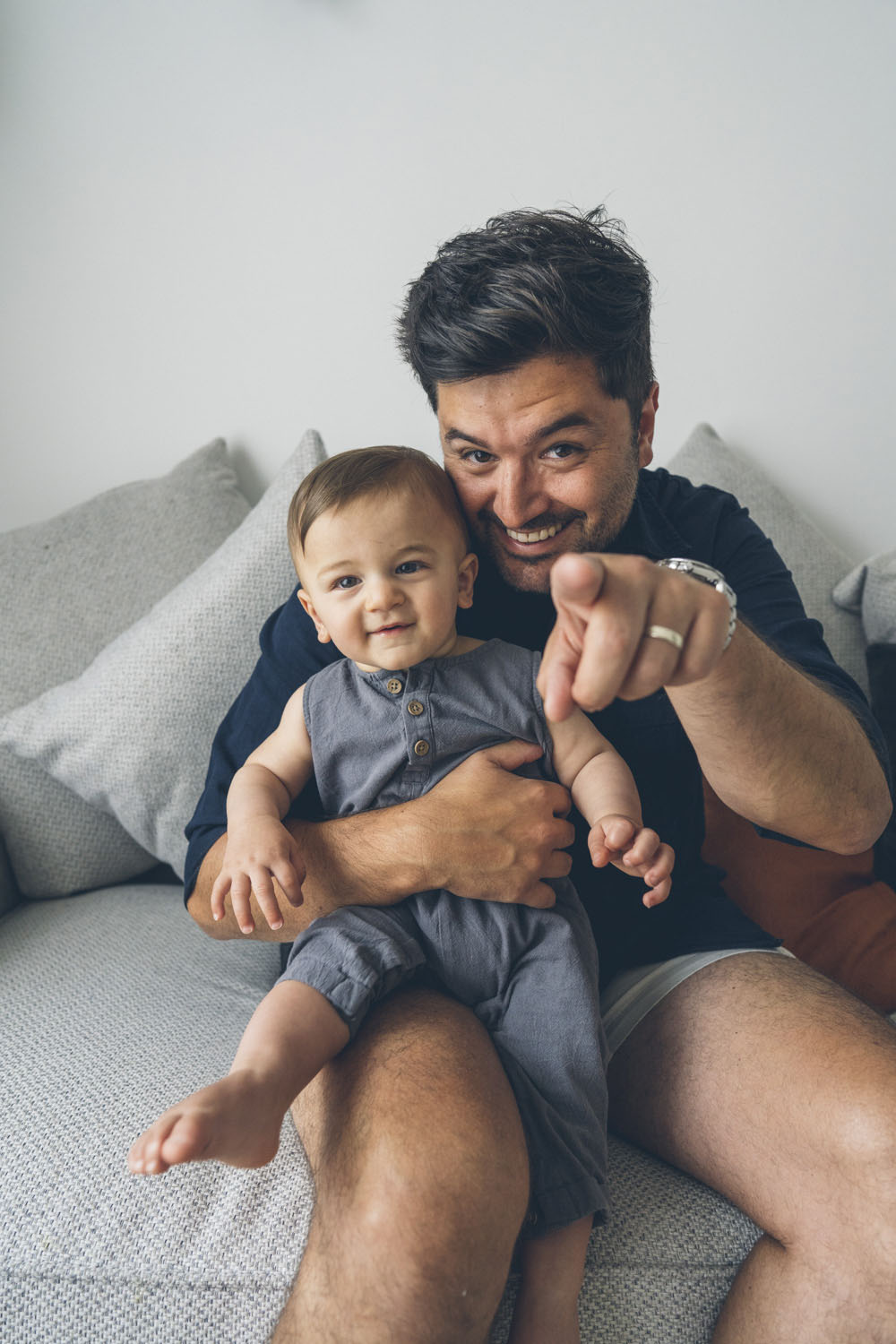 Father sitting on a couch holding his baby son and pointing at the camera laughing during an in-home documentary family photography session in Melbourne