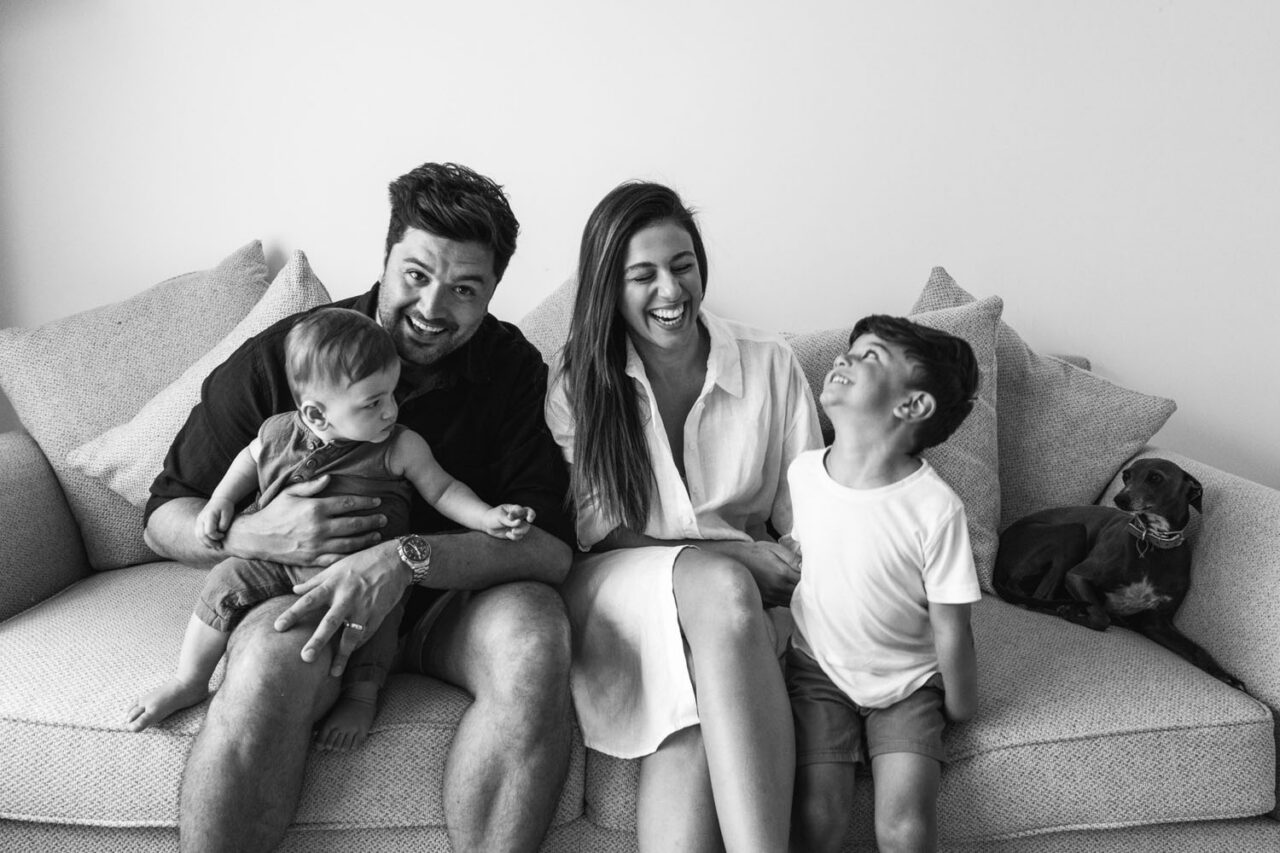 Family of four sitting on a couch laughing together during an in-home documentary family session in Melbourne, with a small dachshund tucked in at the end of the couch