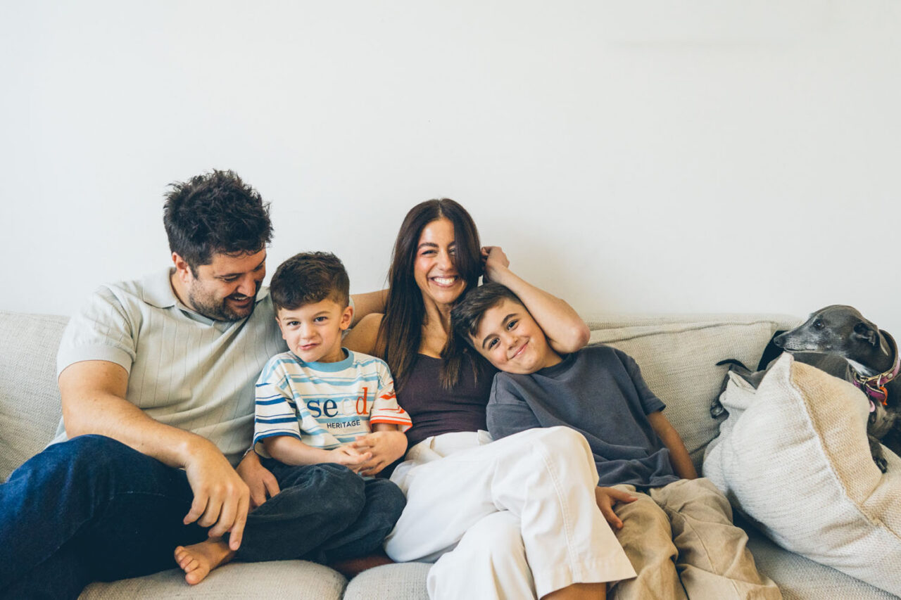 Family of four sitting together on a couch smiling during an in-home documentary Your Story session in Melbourne, Italian greyhound dog perched at the end of the couch