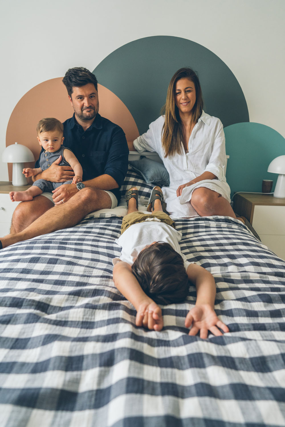 Family of four sitting on a bed during an in-home documentary family photography session in Melbourne, toddler lying face down on the bed in the foreground while parents sit calmly with baby behind him