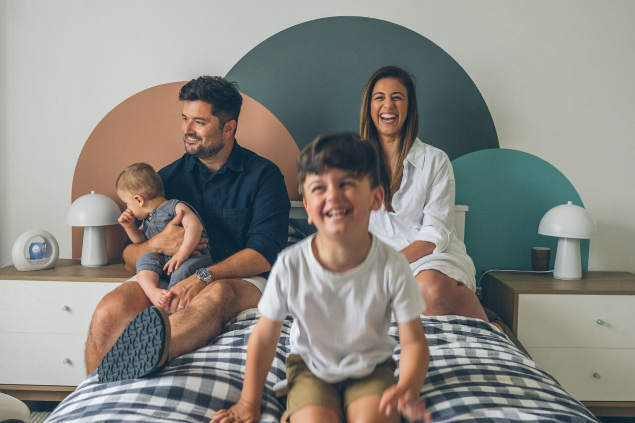 Family of four sitting on a bed laughing during an in-home documentary family photography session in Melbourne, toddler in the foreground grinning at camera