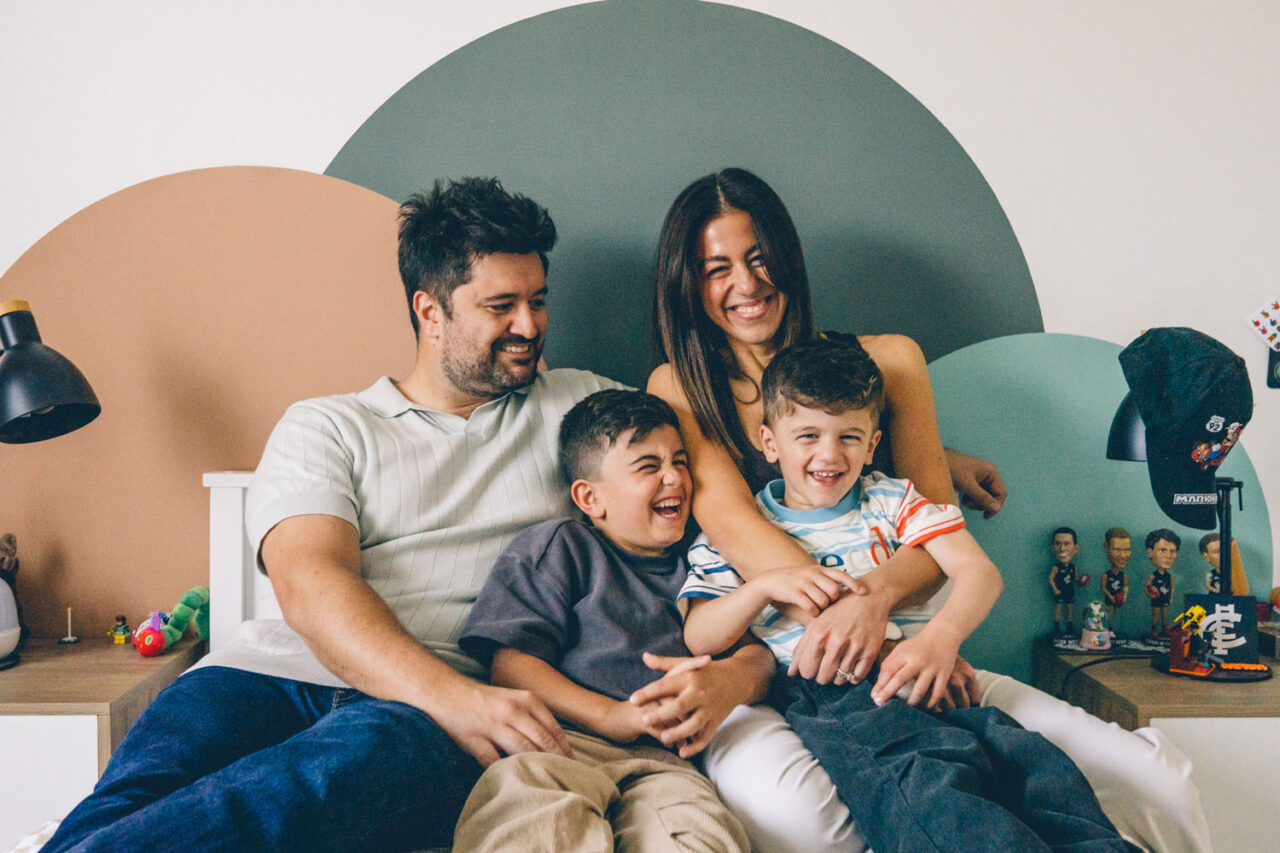 Family of four sitting together on a bed laughing in a colourful painted bedroom during an in-home documentary Your Story session in Melbourne, two boys giggling between their parents