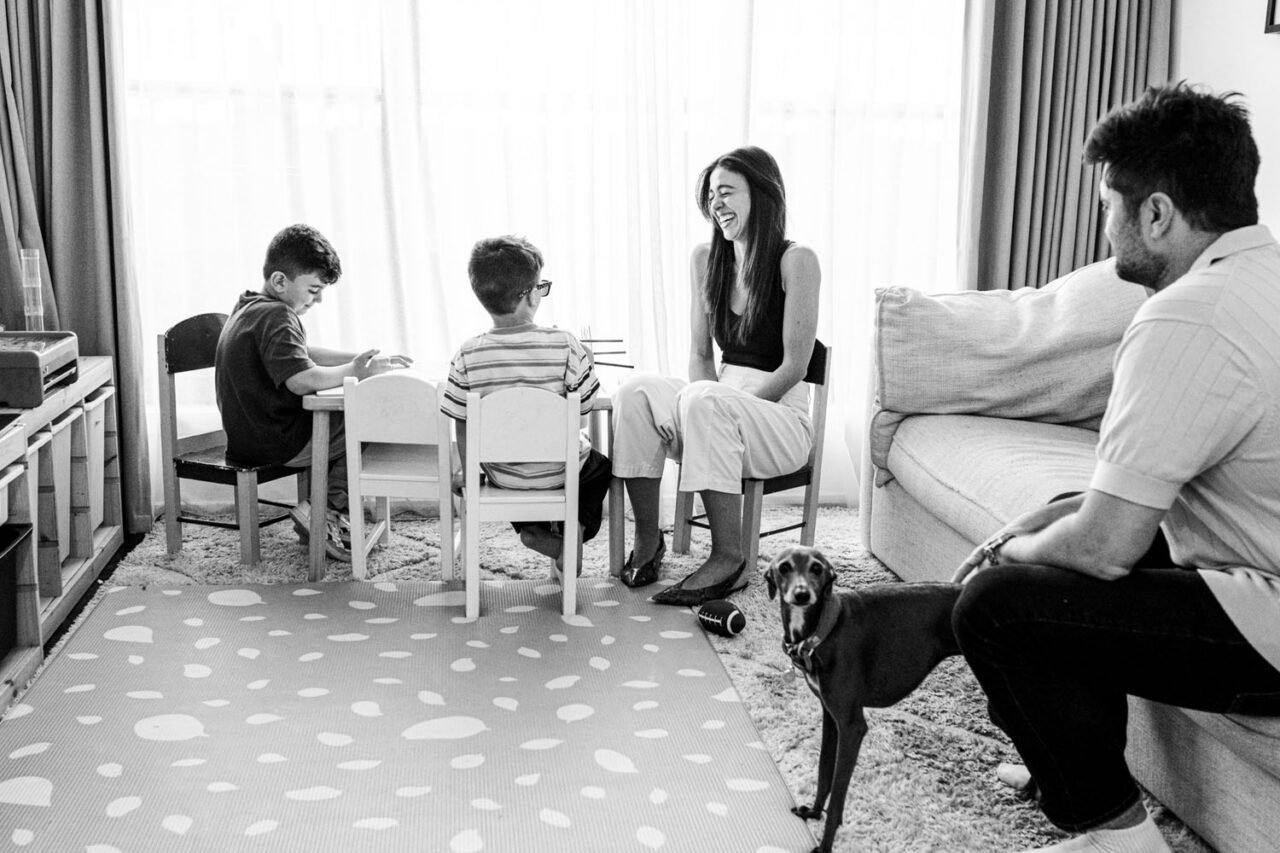 Family sitting together in a playroom during an in-home documentary Your Story session in Melbourne, two boys at a small table, mother laughing, father watching, small dark dog in the foreground