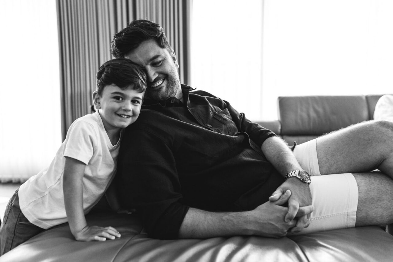 Father and young son sitting together on a couch laughing during an in-home documentary family session in Melbourne