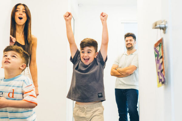 Family of four playing basketball in the hallway of their Melbourne home during a Your Story documentary family session, older boy celebrating with arms raised, younger boy holding the ball, mother laughing and father watching on with arms crossed