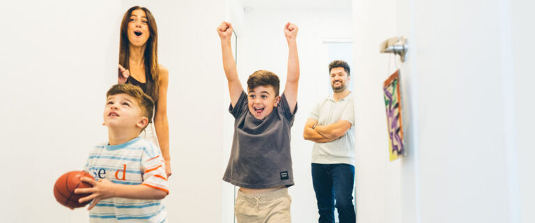 Family of four playing basketball in the hallway of their Melbourne home during a Your Story documentary family session, older boy celebrating with arms raised, younger boy holding the ball, mother laughing and father watching on with arms crossed