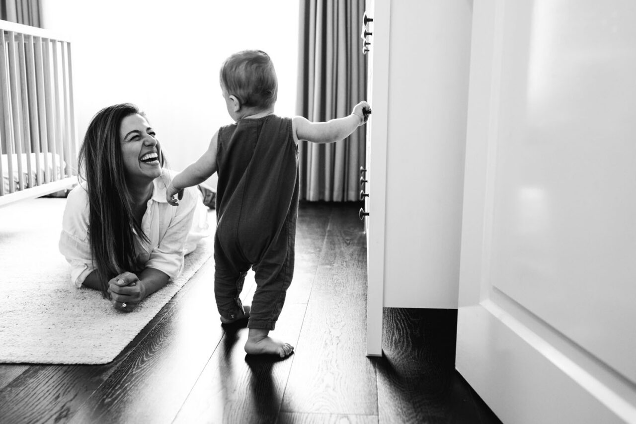 Mother lying on the floor laughing while her baby stands and reaches for a door handle during an in-home documentary family session in Melbourne
