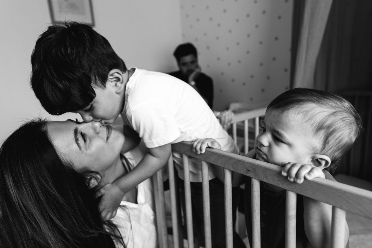 Mother receiving a kiss from her toddler son leaning over a cot while baby looks on, father softly out of focus in the background, documentary family session Melbourne