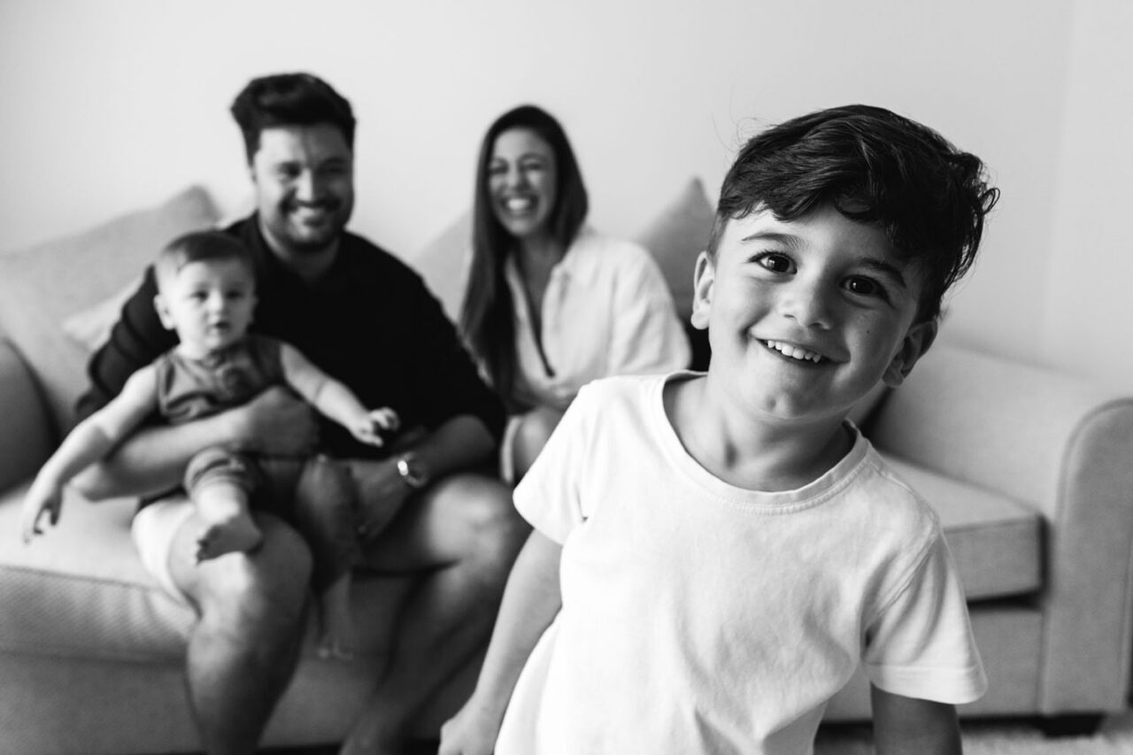 Cheeky toddler boy grinning at camera in the foreground with both parents and baby sibling smiling softly on the couch behind him during an in-home documentary family session in Melbourne