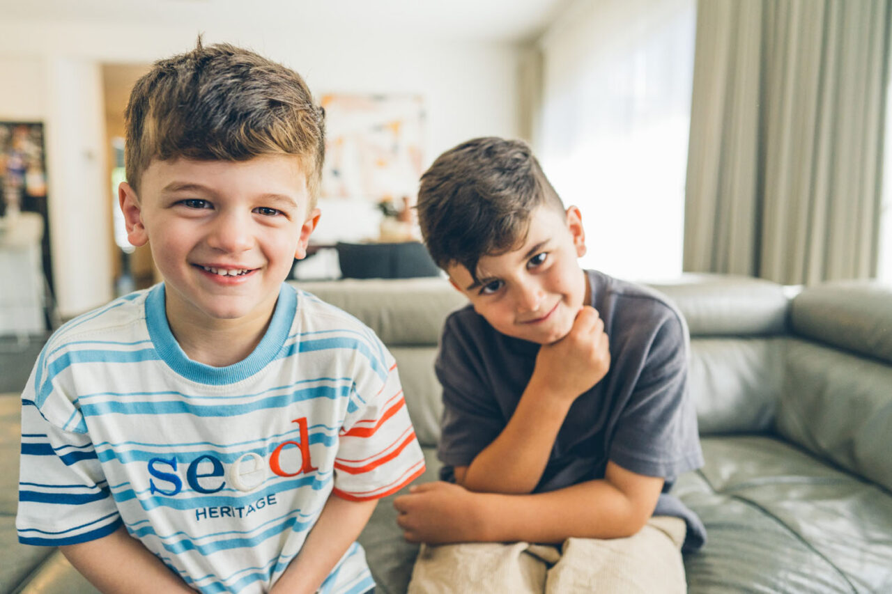 wo brothers sitting together on a couch looking at the camera during an in-home documentary family session in Melbourne, one grinning widely and one with a quiet knowing smile