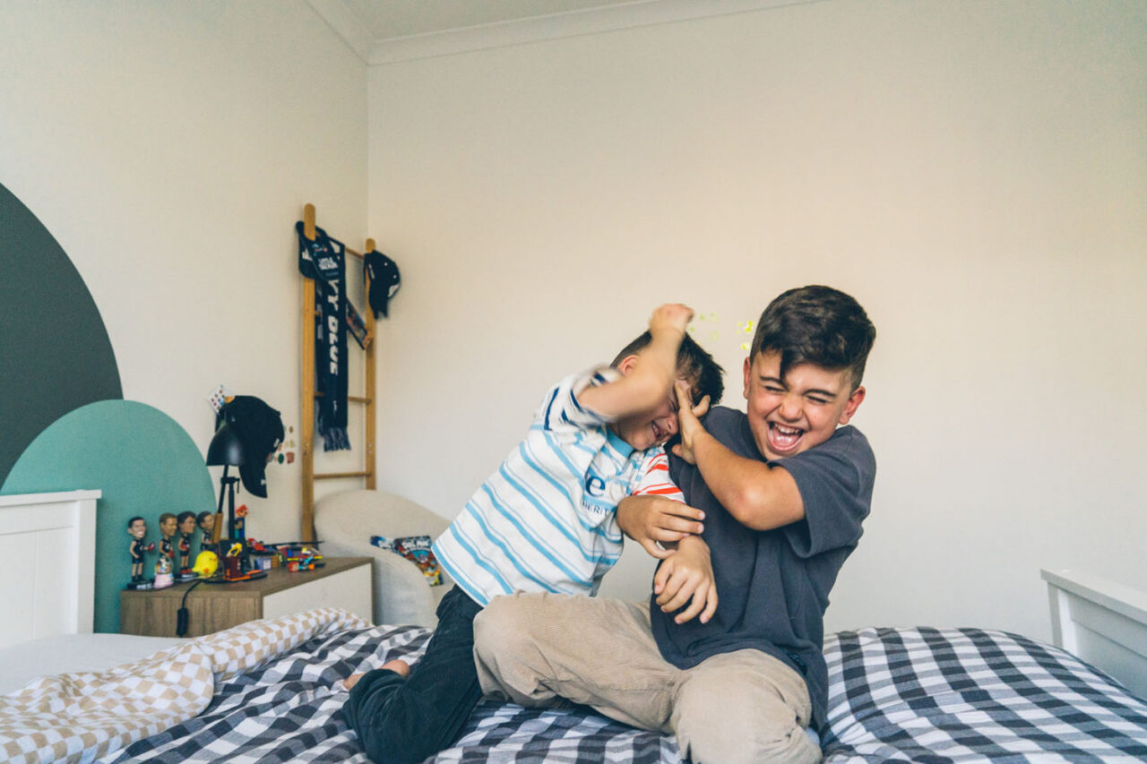 Two brothers wrestling and laughing together on a bed during an in-home documentary family session in Melbourne, younger boy grabbing older brother who is laughing with his whole body