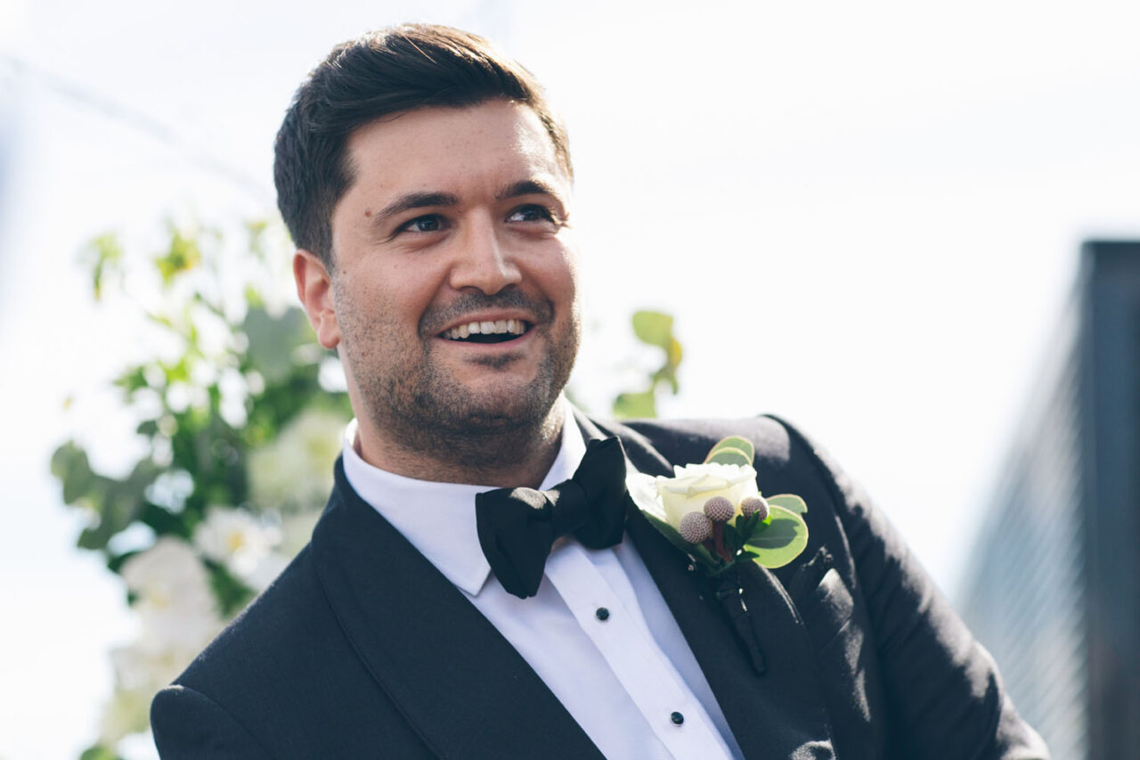 Groom smiling during outdoor wedding ceremony at Luminare South Melbourne, wearing black tuxedo with white rose boutonniere