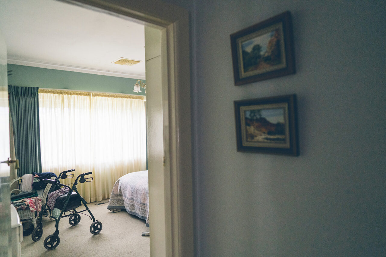 View through a doorway into a bedroom with a walking frame and framed paintings on the wall, documentary family home photography Melbourne