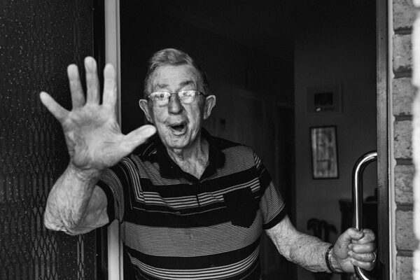 Black and white photograph of an elderly man waving from his front door saying bye, documentary family home photography Melbourne