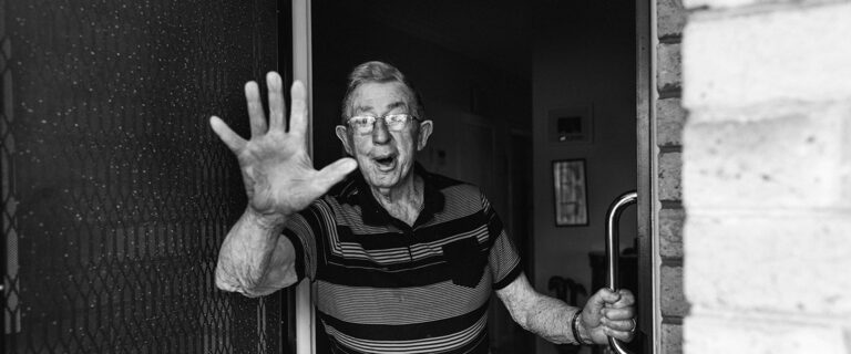 Black and white photograph of an elderly man waving from his front door saying bye, documentary family home photography Melbourne