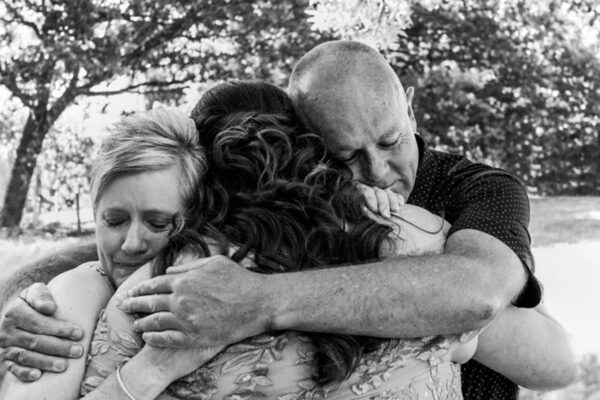 Black and white documentary photograph of a bride being embraced by both parents in an emotional three way hug at an LGBTQI wedding, captured by LJM Photography Melbourne