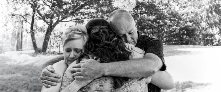 Black and white documentary photograph of a bride being embraced by both parents in an emotional three way hug at an LGBTQI wedding, captured by LJM Photography Melbourne