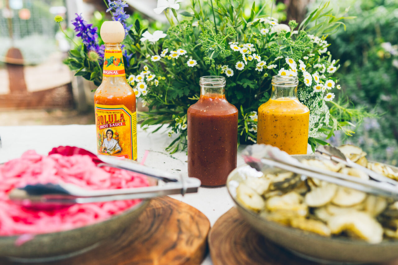 Armstrong BBQ condiment table at a backyard wedding with Cholula hot sauce, homemade BBQ sauces and pickled vegetables surrounded by homegrown flowers
