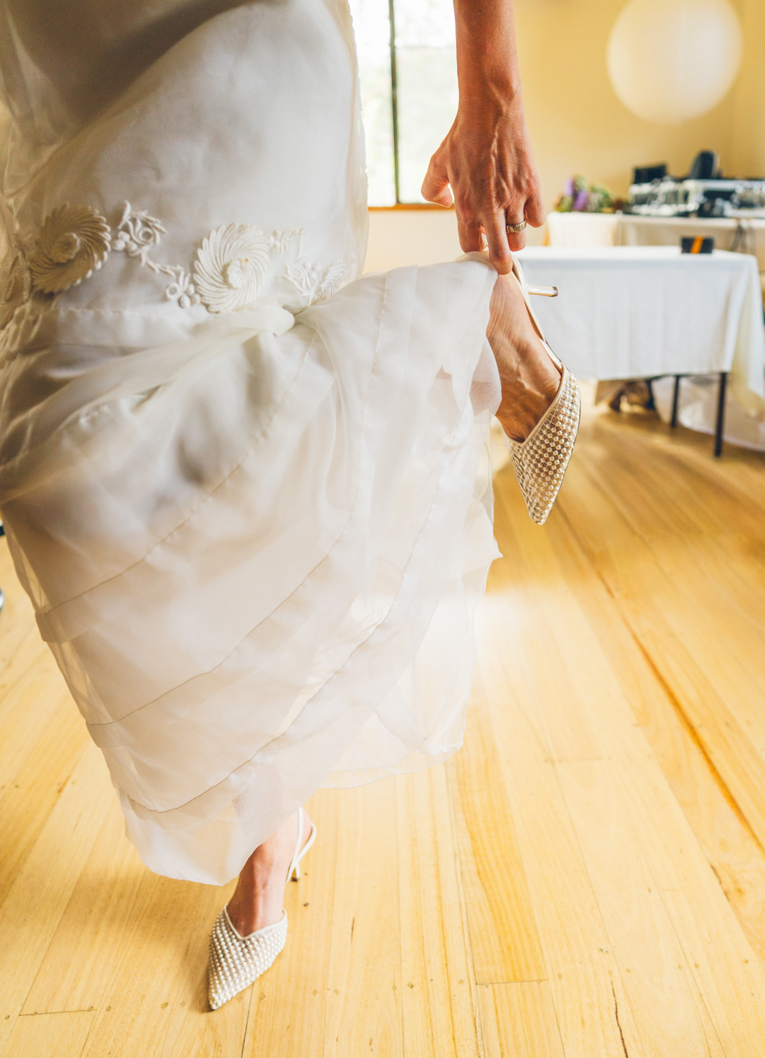 Close up of a bride walking in a layered white vintage wedding dress and pearl-studded heels, holding her shoe