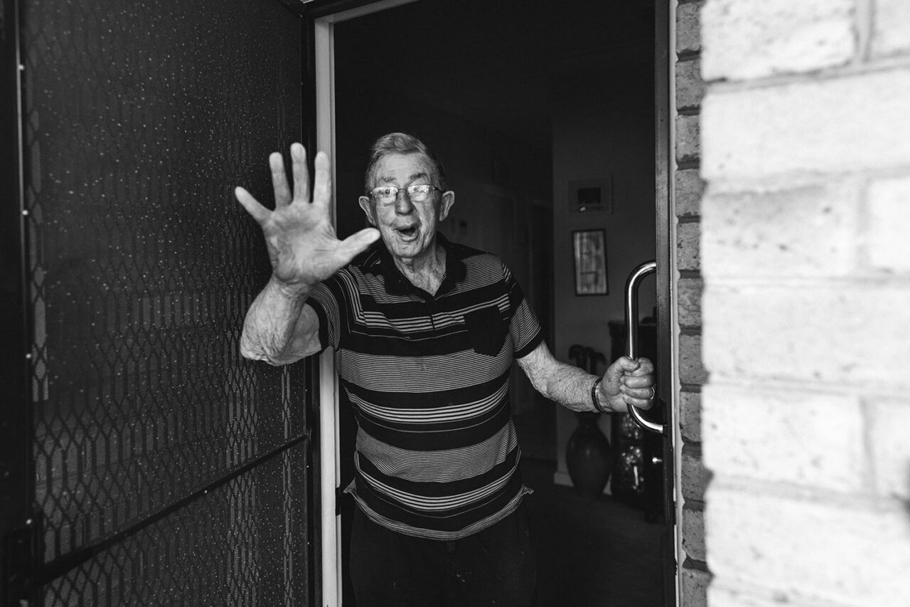 Black and white documentary portrait of an elderly man opening his front door with his hand raised in surprise and delight, Your Story session by LJM Photography Melbourne