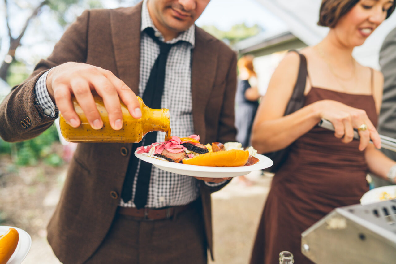 Wedding guest in a brown suit pouring mustard sauce over a plate of American BBQ brisket at a backyard wedding reception