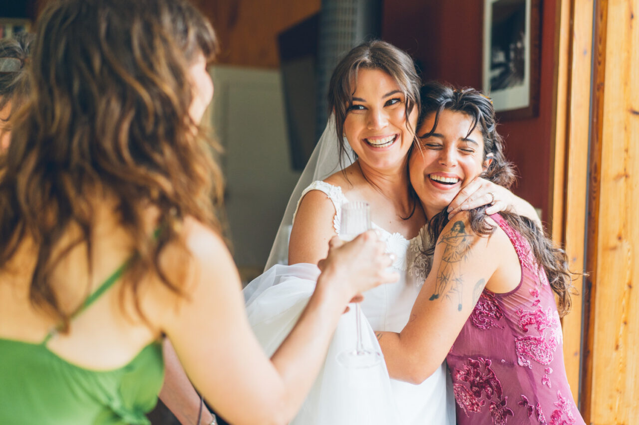 Bride laughing and embracing a friend in a pink embroidered dress while getting ready, with another friend smiling in the background