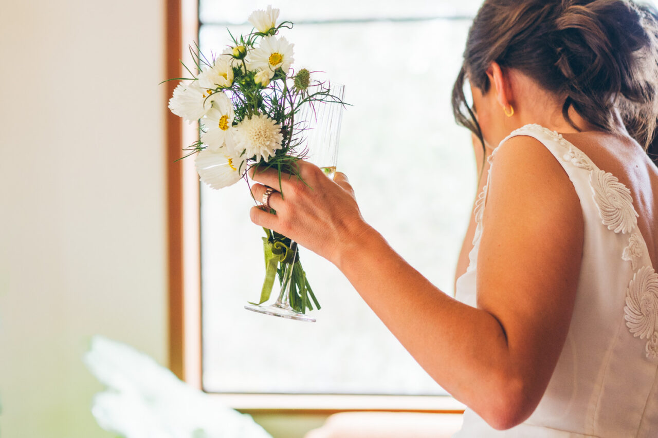 Bride in a vintage white wedding dress holding a small homegrown bouquet of white cosmos and daisies with a champagne glass, looking down by a window